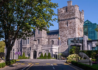 An Exterior landscape shot of Clontarf Castle, a Large tree sits to the left foreground of the shot. The castle has grey brick work, and behind the tower is a new glass lobby area.