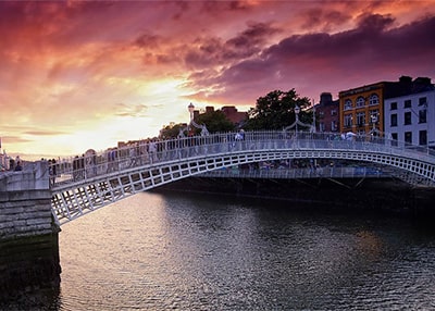 A landscape image of Dublin Bridge, historically known as the Ha'penny Bridge. The cast iron bridge, painted white has a large arch to the middle. Sun sets to the rear of the bridge, with a pink and orange glow to the sky. People are crossing the bridge by foot.