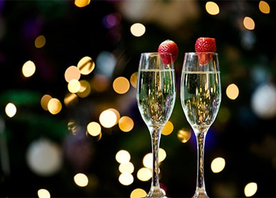 Two filled champagne flutes sit centre focus of the shot. They have a strawberry on the rim. The background is out of focus with fairy lights dotted around the image.