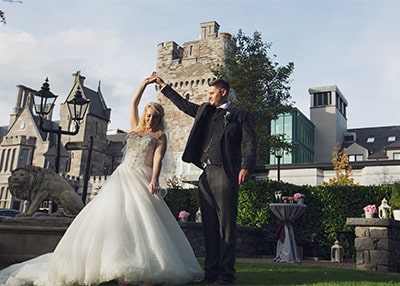 An Exterior landscape shot of Clontarf Castle, a Large tree sits to the left foreground of the shot. The castle has grey brick work, and behind the tower is a new glass lobby area.