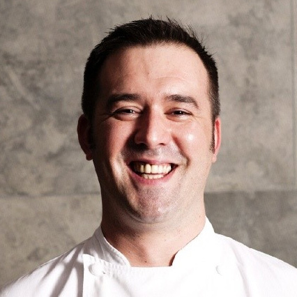 A portrait head shot of Executive Head Chef Paul Devoy. He is smiling looking straight to the camera, contrasting from a grey wall behind him.